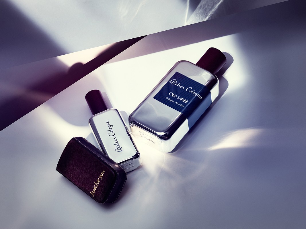 Close-up product shot of two Atelier Cologne bottles, one with a dark leather case, angled on a bright white surface with shadows.