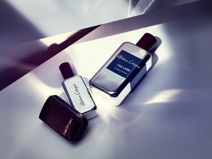 Close-up product shot of two Atelier Cologne bottles, one with a dark leather case, angled on a bright white surface with shadows.