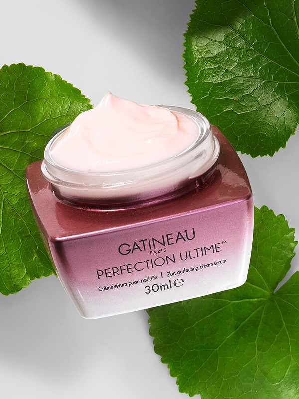 Gatineau Perfection Ultime cream with clear lid, pink cream, surrounded by large green leaves against a white background, close-up.