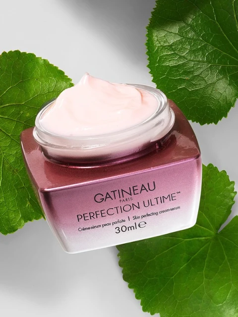 Gatineau Perfection Ultime cream with clear lid, pink cream, surrounded by large green leaves against a white background, close-up.