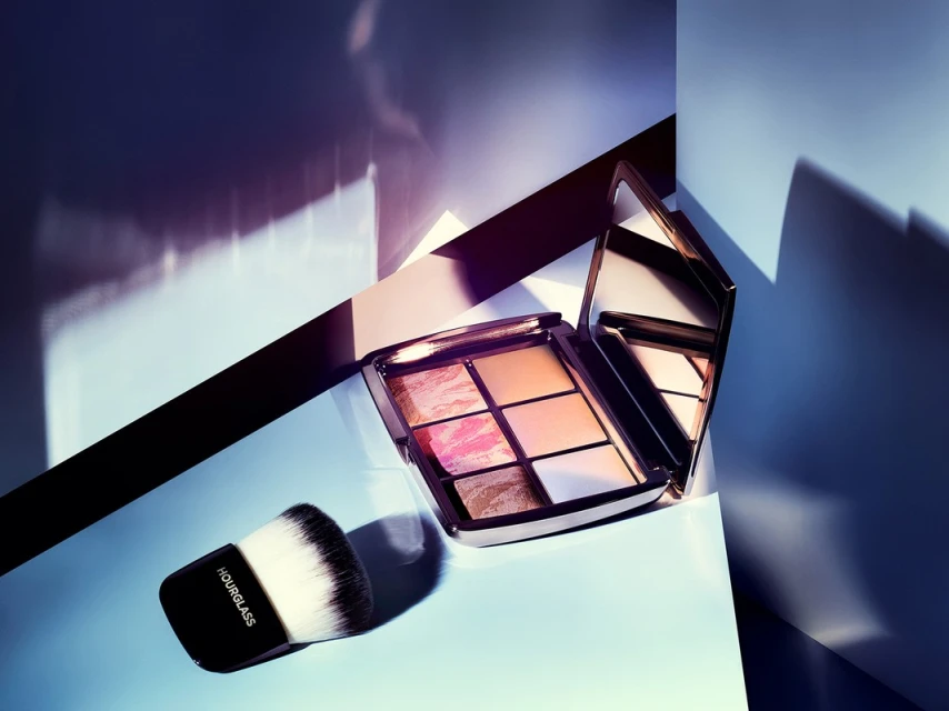 Still life studio shot of an Hourglass Ambient Lighting Palette with mirror and kabuki brush against a blue and purple geometric background.