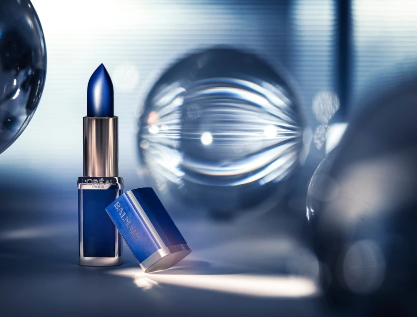 Still life studio shot of blue L'Oreal lipstick with silver and blue packaging, surrounded by glass spheres and blue background lighting.