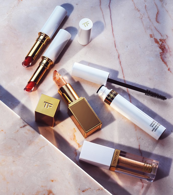 Several tubes of Tom Ford lipstick and mascara are displayed on a marbled pink surface in a cosmetic flat lay studio shot.