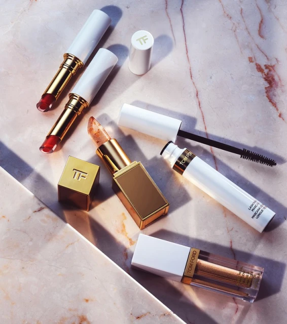Several tubes of Tom Ford lipstick and mascara are displayed on a marbled pink surface in a cosmetic flat lay studio shot.