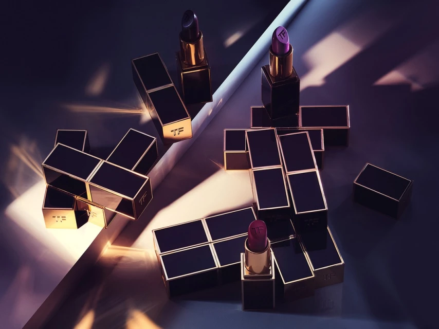 Still life of Tom Ford lipsticks in black and gold cases, arranged geometrically on a surface with linear light and shadow.