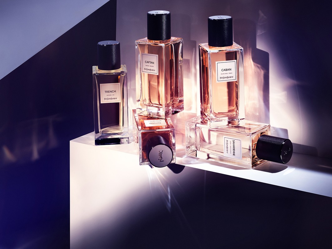 Five glass perfume bottles with black caps arranged on a white podium under dramatic angular lighting.
