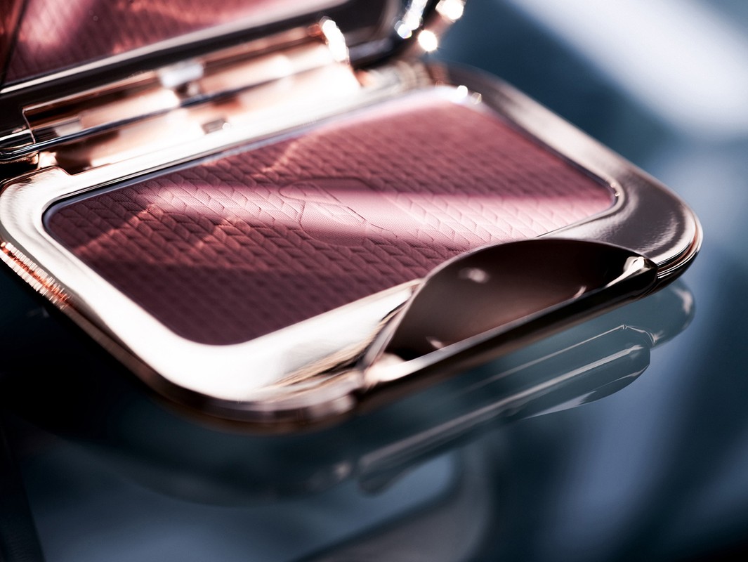 Close-up of an open metallic makeup compact with textured mauve powder product inside on a dark reflective surface.