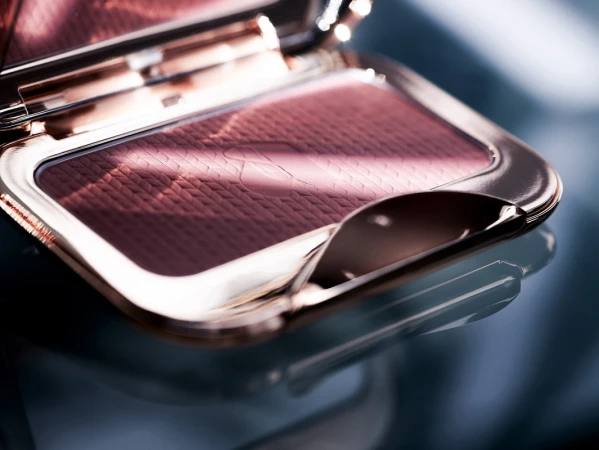 Close-up of an open metallic makeup compact with textured mauve powder product inside on a dark reflective surface.