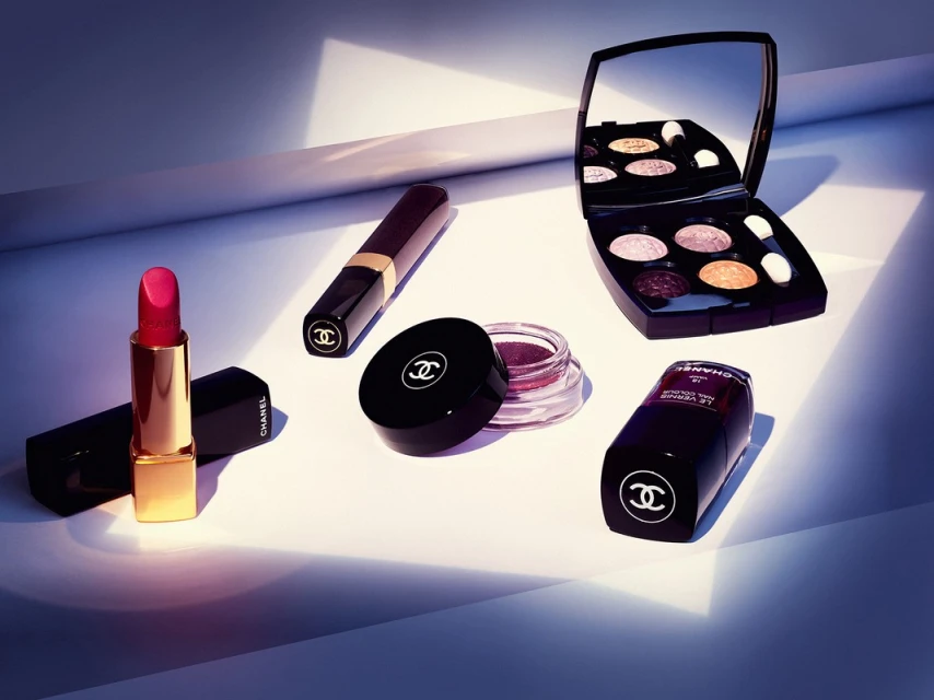 Still life shot of Chanel makeup products including lipstick, eyeshadow, and nail polish, set against a clean, illuminated background.