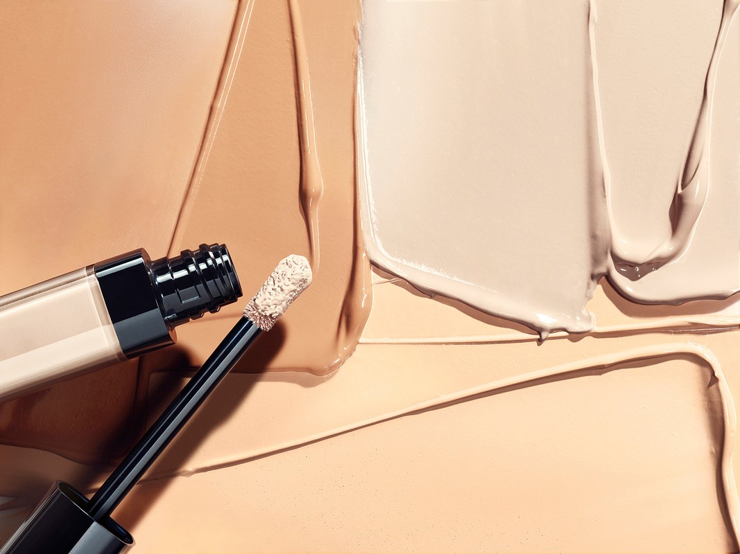 Cosmetic concealer bottle with wand applicator on background of creamy beige foundation and skin tone matching makeup swatches.