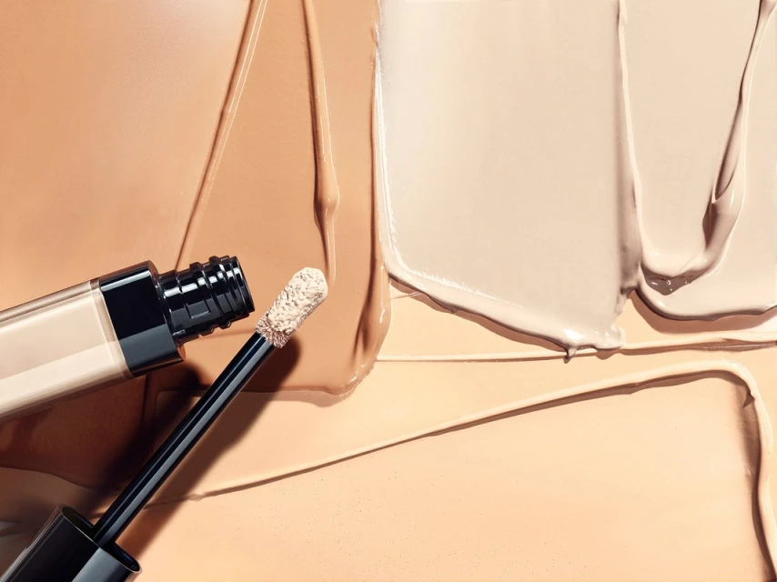Cosmetic concealer bottle with wand applicator on background of creamy beige foundation and skin tone matching makeup swatches.