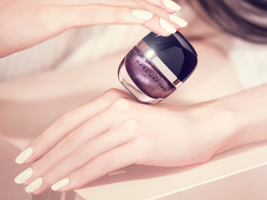 Close-up of pale hands holding a purple glitter Marc Jacobs nail polish bottle, set against a soft pink background in a beauty editorial.
