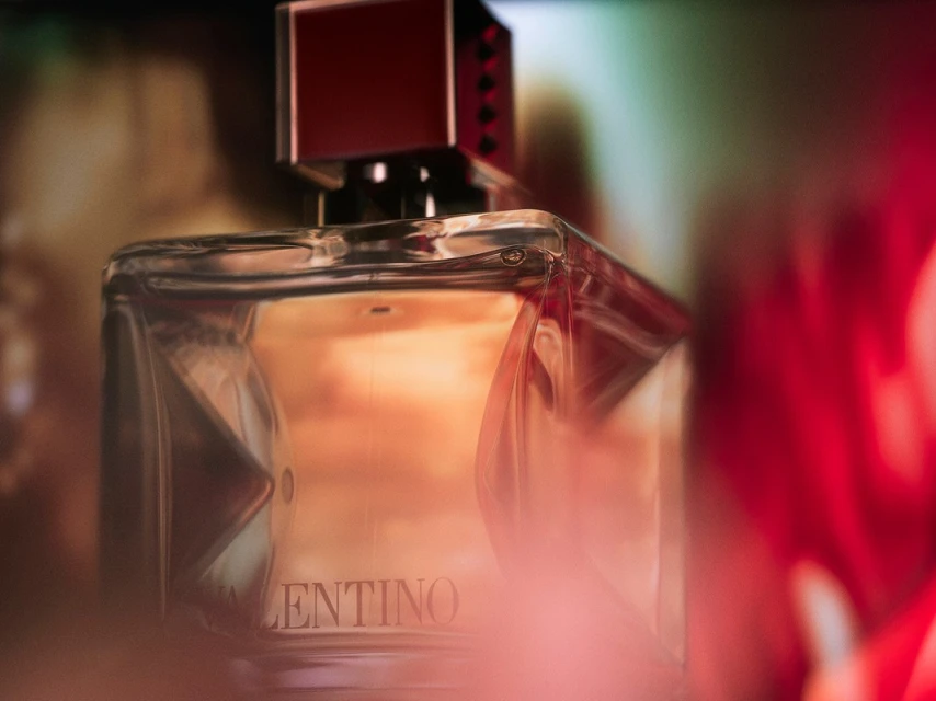 Close-up product shot of a Valentino perfume bottle with a red square cap, set against a blurred background with warm and cool tones.