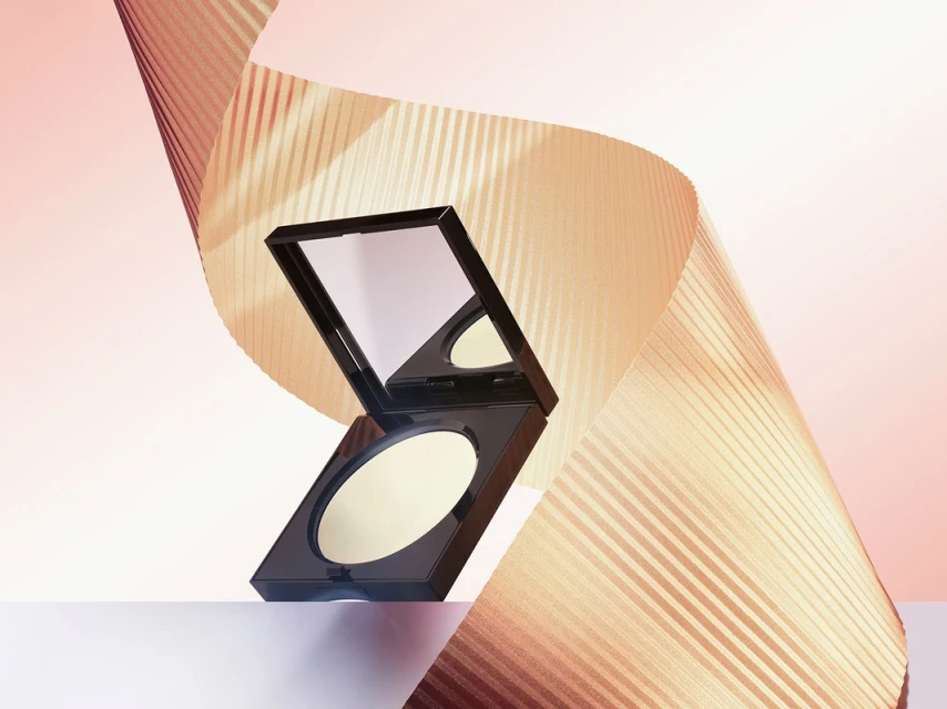 Compact powder foundation case with mirror stands on white surface with swirling textured gold paper in the background, beauty shot.
