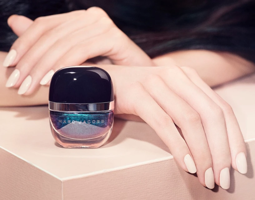 Closeup of a bottle of Marc Jacobs nail polish with a color-shifting finish, set against a cream-colored surface and human hands.