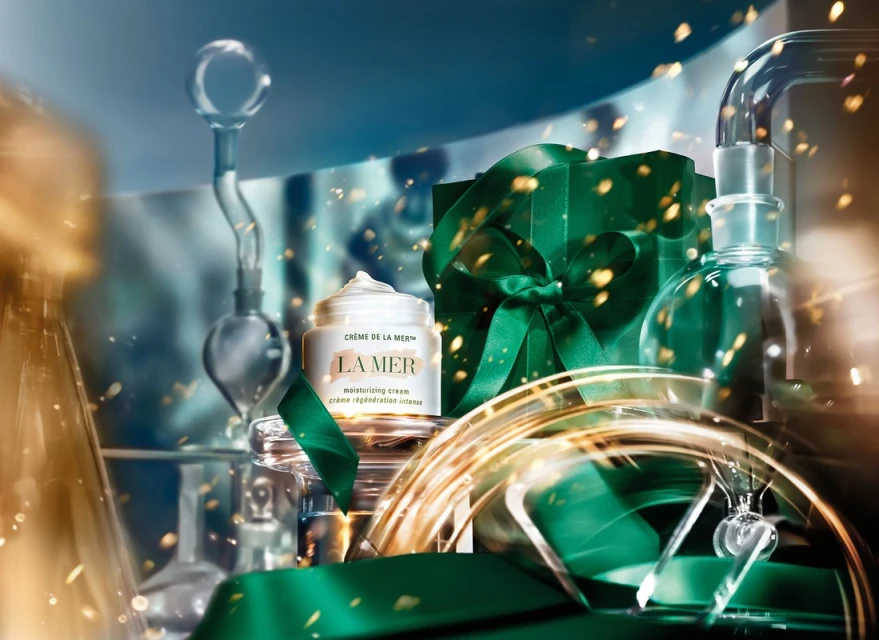 La Mer moisturizing cream jar next to a gift wrapped in green ribbon, and clear laboratory glassware with golden sparkle effects.