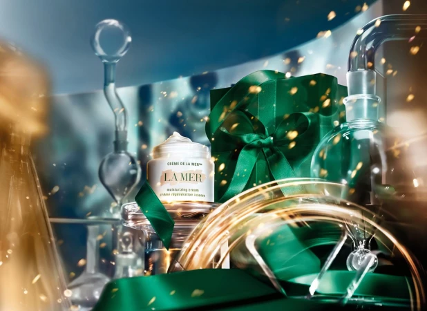 La Mer moisturizing cream jar next to a gift wrapped in green ribbon, and clear laboratory glassware with golden sparkle effects.