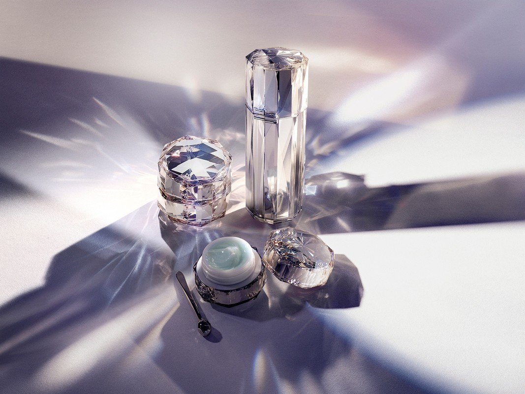 Crystal-faceted cosmetic containers with dispenser, lidded jar, and applicator spoon on a white surface with refracted light patterns.
