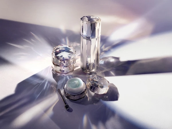 Crystal-faceted cosmetic containers with dispenser, lidded jar, and applicator spoon on a white surface with refracted light patterns.
