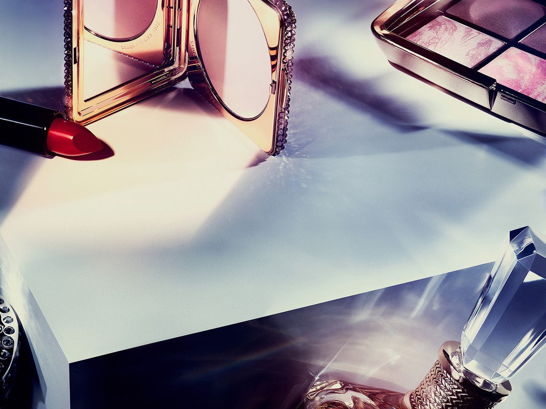An overhead shot displays lipstick, makeup palette, face powder and perfume on a light surface with cast shadows and strong directional lighting.