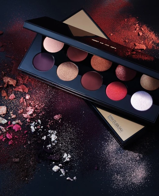 Eye shadow palette in black and gold packaging with a variety of powder colors, and scattered powder on a black background.