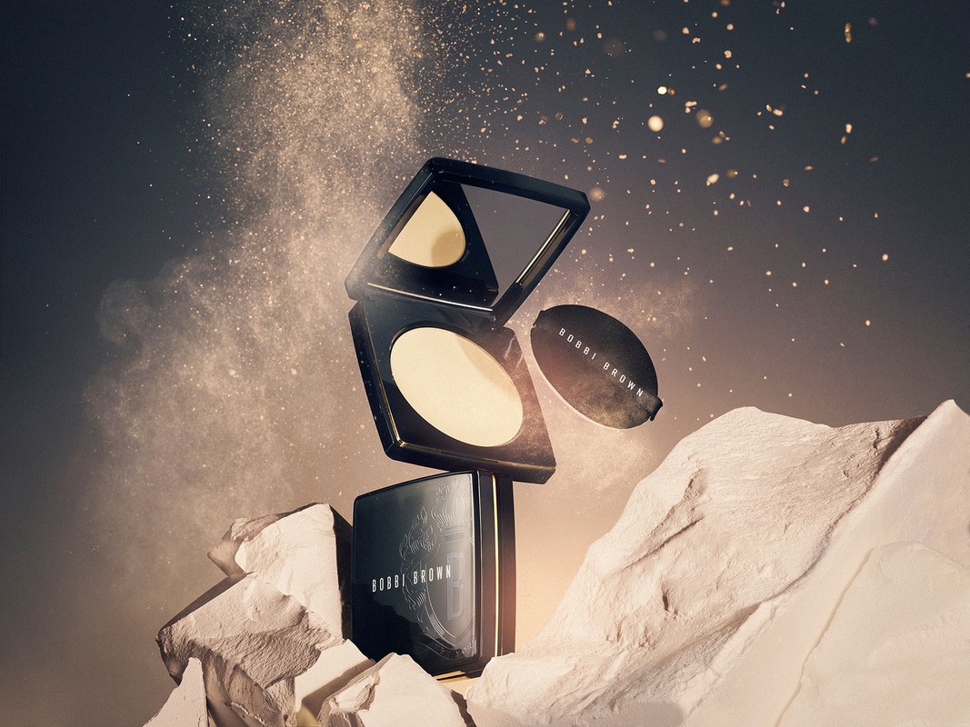 Bobbi Brown pressed powder compact with powder puff on a textured white rock surface with cosmetic powder floating in the air.
