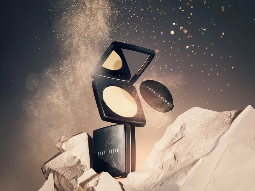 Bobbi Brown pressed powder compact with powder puff on a textured white rock surface with cosmetic powder floating in the air.