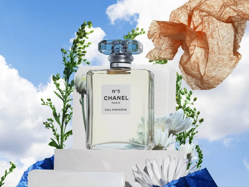 Chanel N°5 Eau Premiere perfume bottle on white blocks with white daisy and green sprigs, set against a blue sky and clouds.