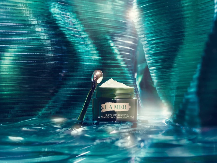 A jar of La Mer eye concentrate cream sits with a silver spoon against an abstract green background for a beauty advertisement.