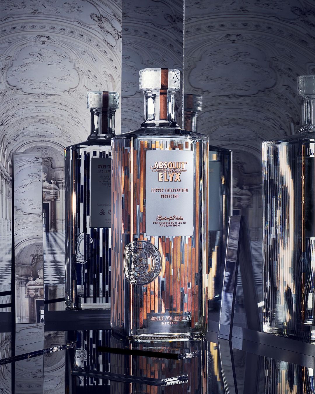 Three crystal bottles of Absolut Elyx vodka on mirrored shelves, illuminated by soft lighting, showcase copper accents and elegant design.