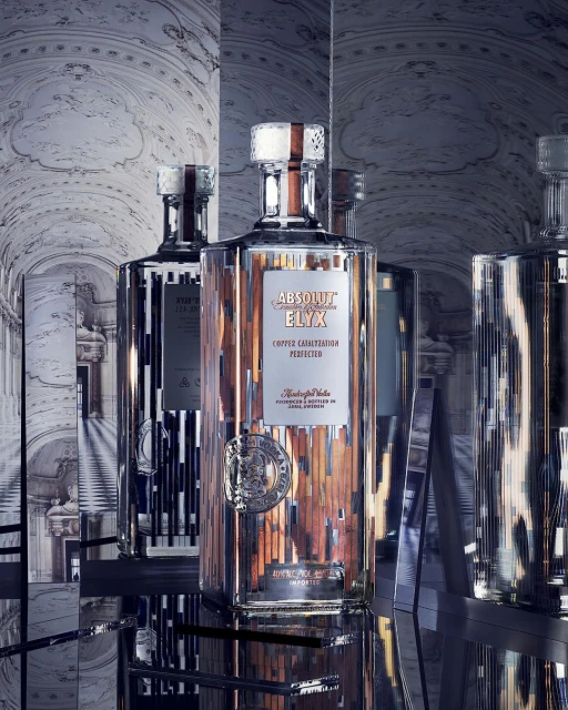 Three crystal bottles of Absolut Elyx vodka on mirrored shelves, illuminated by soft lighting, showcase copper accents and elegant design.