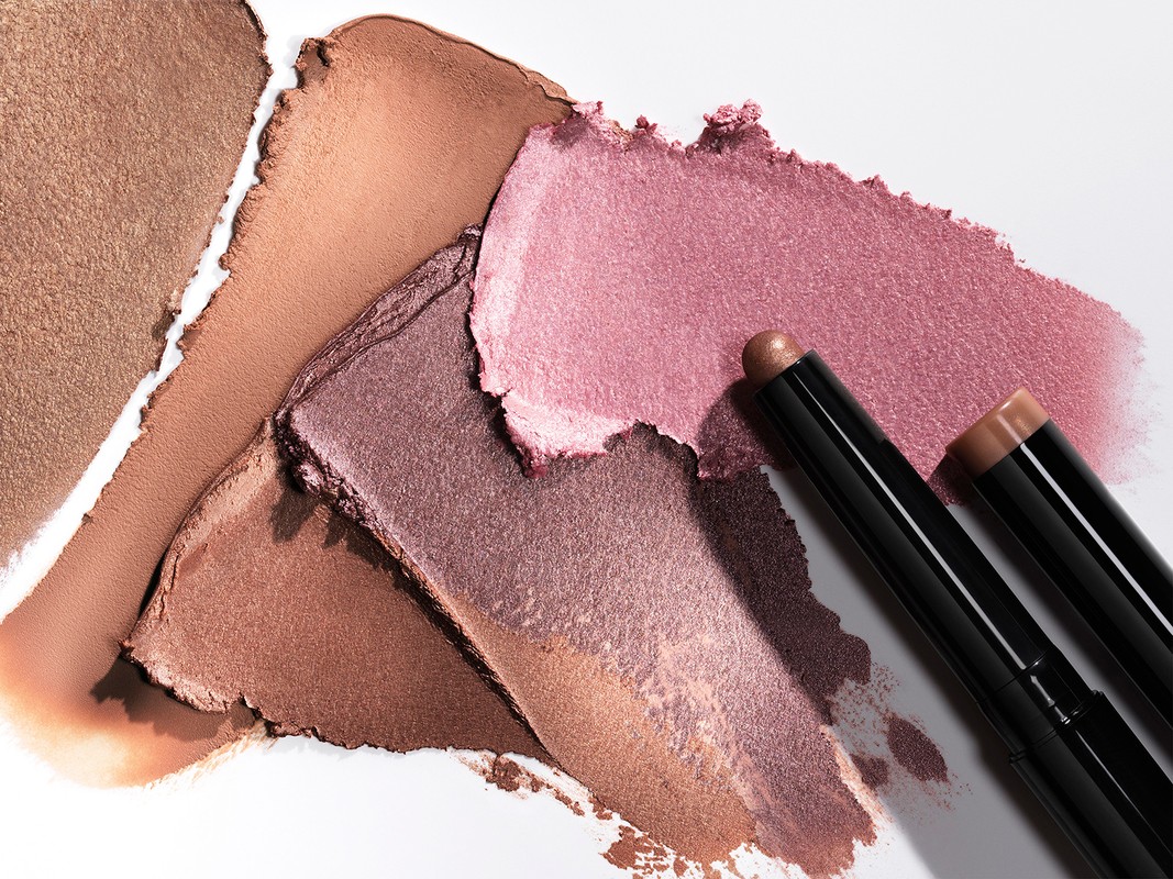 Close-up of cosmetic eye shadow sticks and swatches. Makeup in bronze, brown, and pink tones on a white background in studio lighting.
