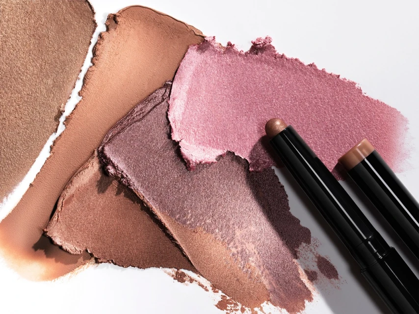 Close-up of cosmetic eye shadow sticks and swatches. Makeup in bronze, brown, and pink tones on a white background in studio lighting.