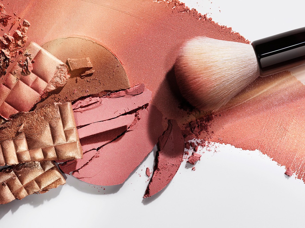 High-angle shot of a makeup brush resting on broken makeup powder, featuring bronze, tan, and peach tones on a white surface.