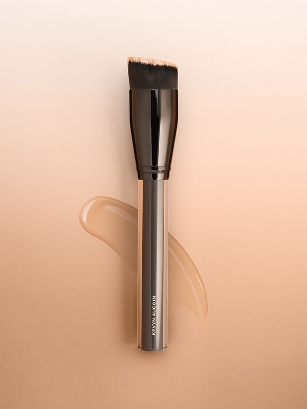 Angled foundation brush by Kevyn Aucoin set against a light gradient background with a swatch of foundation.