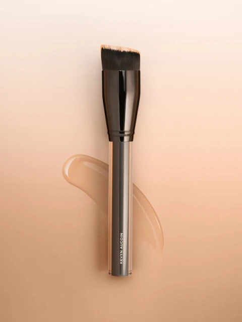Angled foundation brush by Kevyn Aucoin set against a light gradient background with a swatch of foundation.