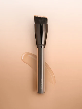 Angled foundation brush by Kevyn Aucoin set against a light gradient background with a swatch of foundation.