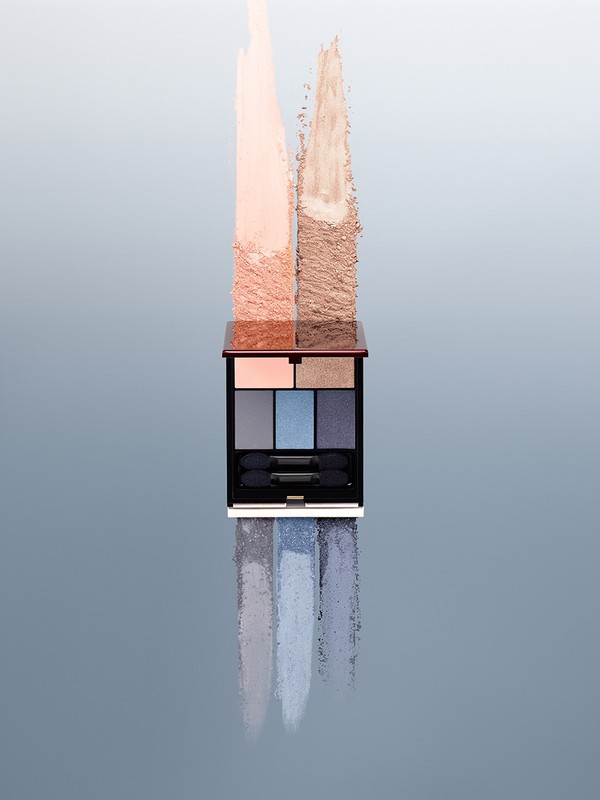 Eye shadow palette with swatches in neutral and blue tones, makeup product shot on a reflective surface against a gradient backdrop.