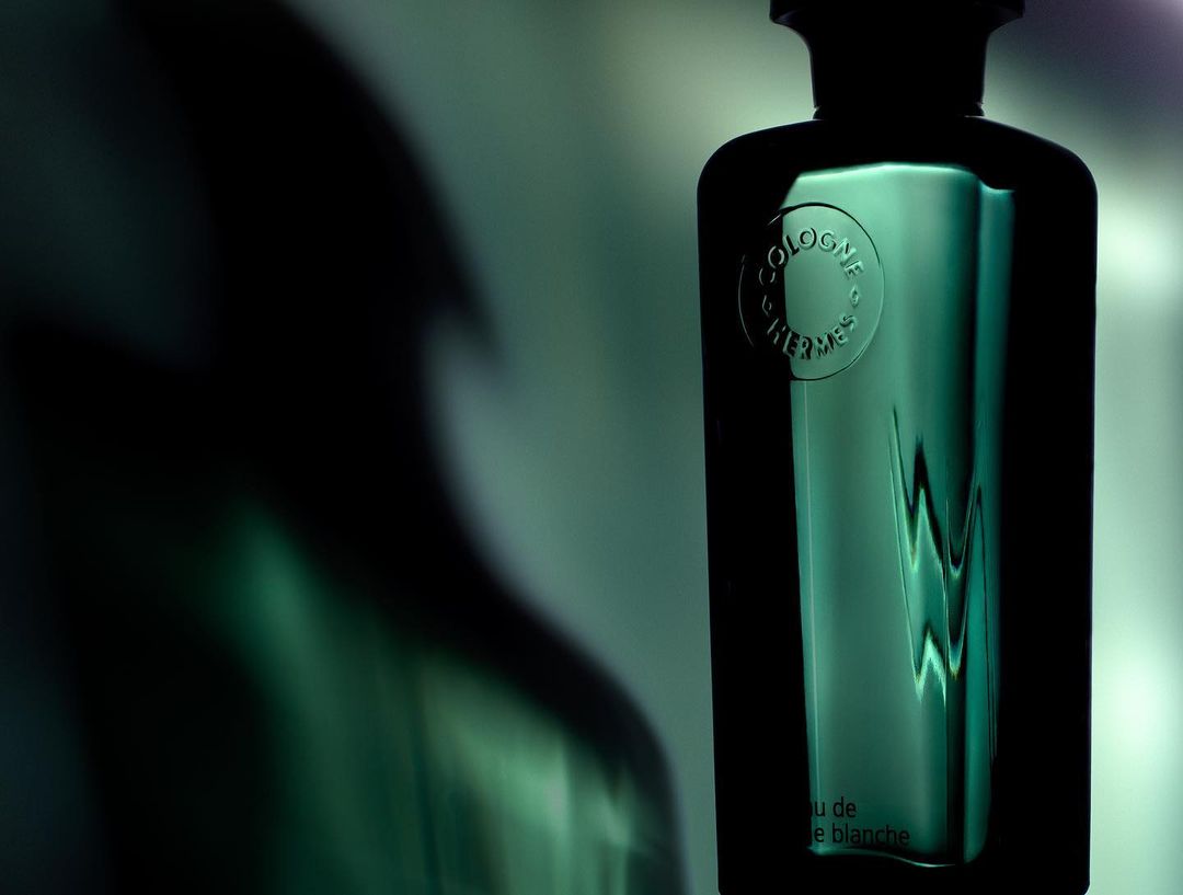 Elegant green perfume bottle from Cologne Hermes stands out against a blurred, shadowy background, highlighting its sleek design and embossed logo.