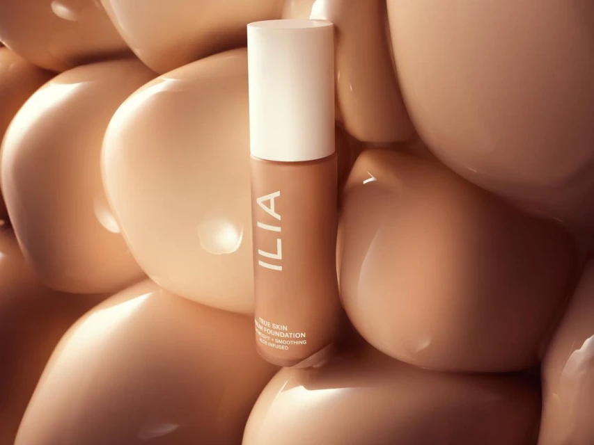 Close-up of Ilia True Skin Serum Foundation bottle against a bubbly, tan-colored background, showcasing beauty product in a studio setting.