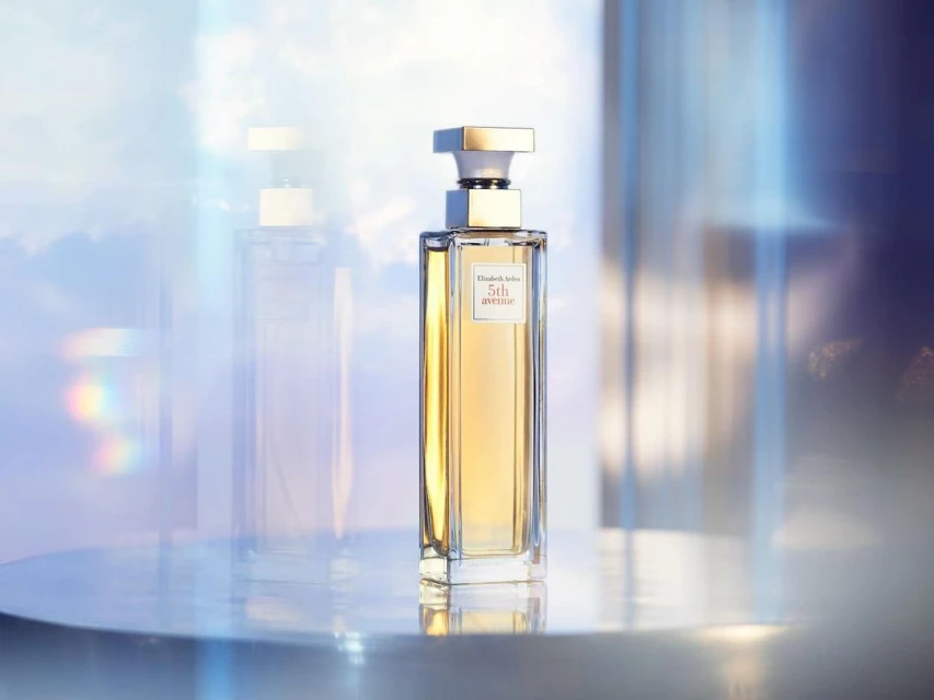 Elizabeth Arden 5th Avenue perfume bottle on a reflective surface with a blue and white bokeh background in a beauty still life.