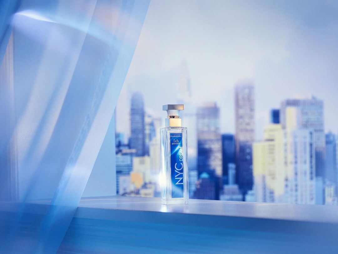 Elizabeth Arden NYC Dreams perfume bottle sits on a ledge with a cityscape backdrop and blue curtain accent, product shot.