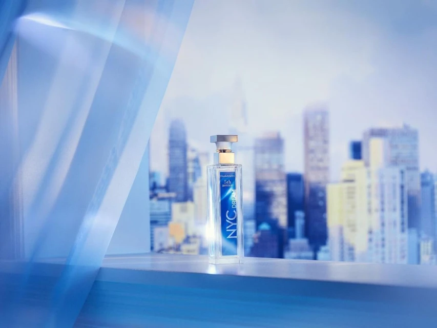 Elizabeth Arden NYC Dreams perfume bottle sits on a ledge with a cityscape backdrop and blue curtain accent, product shot.