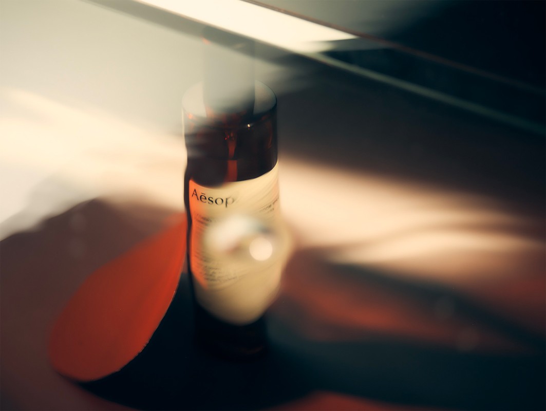 A bottle of Aesop hand balm sits on a surface with light shining through a window, casting shadows across the bottle and surrounding area.