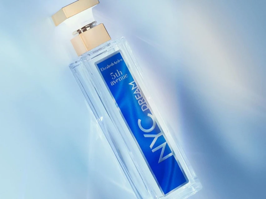 A clear glass Elizabeth Arden 5th Avenue NYC Dream perfume bottle with a gold cap is featured against a light blue gradient background.