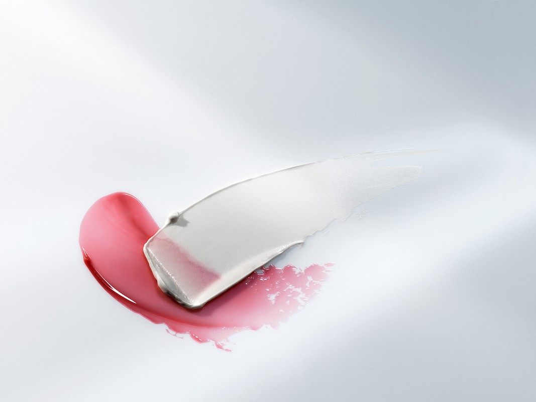 Swatches of pink lip gloss and white cream on a white background with soft lighting, showcasing beauty product textures and application.