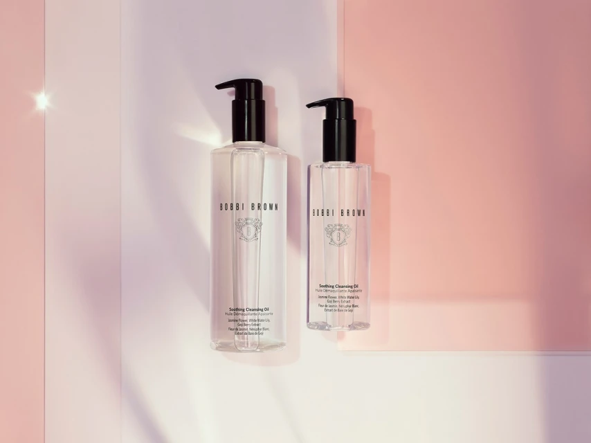 Two bottles of Bobbi Brown Soothing Cleansing Oil are staged against a pink-and-white backdrop, softly lit to showcase the skincare product.