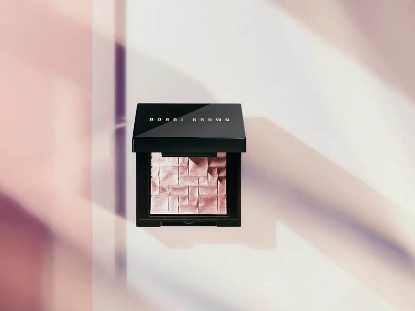 Bobbi Brown highlighting powder, black compact case, and soft pink powder with brick pattern sits against pastel-toned background with shadows.
