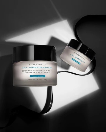 Two jars of Skinceuticals A.G.E. Interrupter Advanced cream sit with a black ribbon on a surface in a studio with artificial lighting.