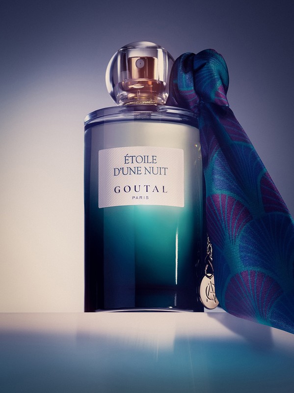 Close-up of an Etoile D'une Nuit Goutal Paris perfume bottle next to a patterned turquoise and purple silk scarf on a reflective surface.